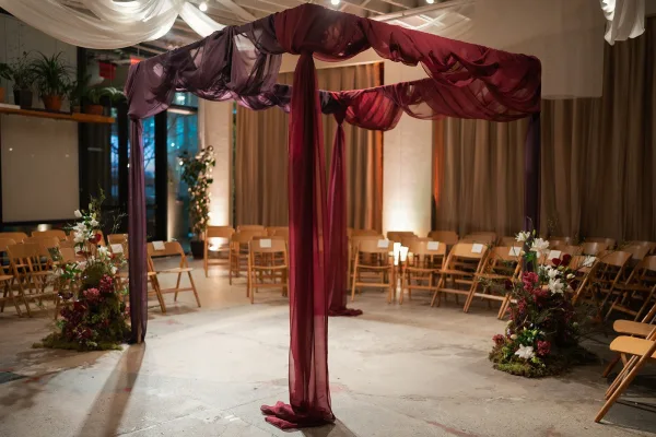 Ceremony setup with a fabric draped wedding arch, sheer canopy and floral arrangements, with wood folding chairs in a windowed industrial venue