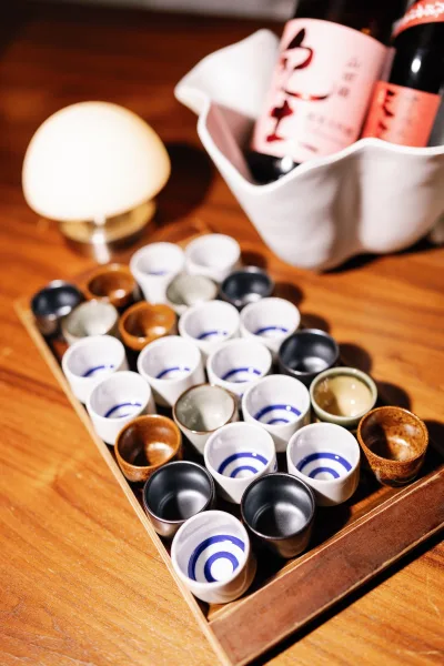 Sake tasting station wedding sake bar with ceramic sake cups on a wooden tray, bottles in an ice bucket, and warm lamp light on a wood table