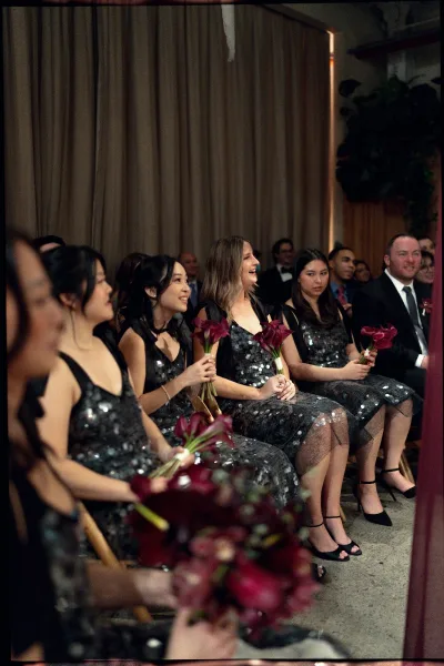 Bridesmaid ceremony seating with black sequin dresses, holding burgundy calla lily bouquets as guests watch in a draped indoor venue
