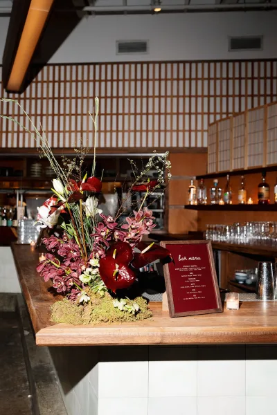 Wedding bar decor with a framed bar menu sign beside anthurium and orchid florals, bottles and glassware on a wood counter under an industrial ceiling