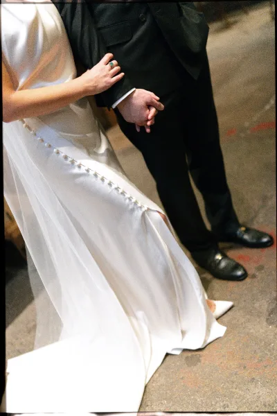 Ceremony moment of bride and groom walking arm in arm, close-up of her button-back gown and emerald engagement ring on a neutral indoor floor