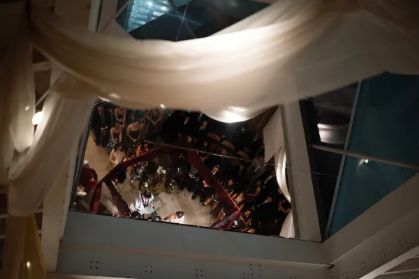 Wedding ceremony overhead shot of couple under a fabric-draped chuppah with floral arrangement, guests seated in a glass-ceiling atrium