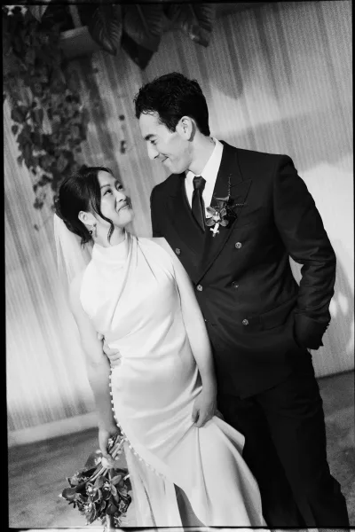 Couple portrait in black and white wedding portrait style, bride and groom looking at each other as she holds a cascading bouquet before draped fabric and greenery backdrop