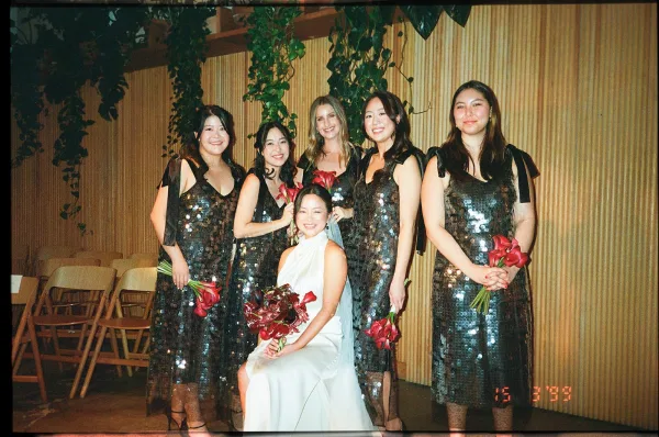 Bridesmaids portrait with bride seated as bridesmaids holding bouquets in sequin dresses before a wood slat wall with hanging greenery