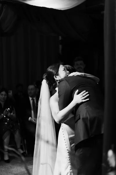 First dance as bride and groom slow dance in close embrace, her button back wedding dress and veil flowing in an indoor reception with guests