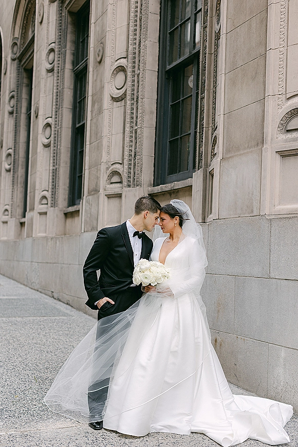 Couple portrait of bride and groom portrait on a city sidewalk, her veil blowing as she holds a white bouquet beside his black tuxedo