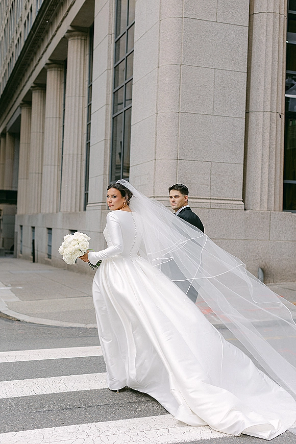 Couple portrait of bride and groom street photo with bride looking back, cathedral veil trailing as they cross a city street by stone columns