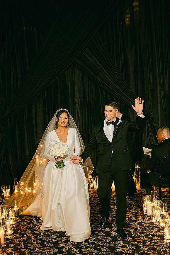 Wedding recessional as bride and groom walk hand in hand down a candlelit aisle, groom waving beside her veil and bouquet amid black drapery