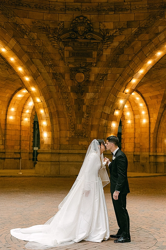 Wedding kiss portrait of bride and groom kissing under a veil, holding hands in a brick courtyard by a stone arch with warm lights