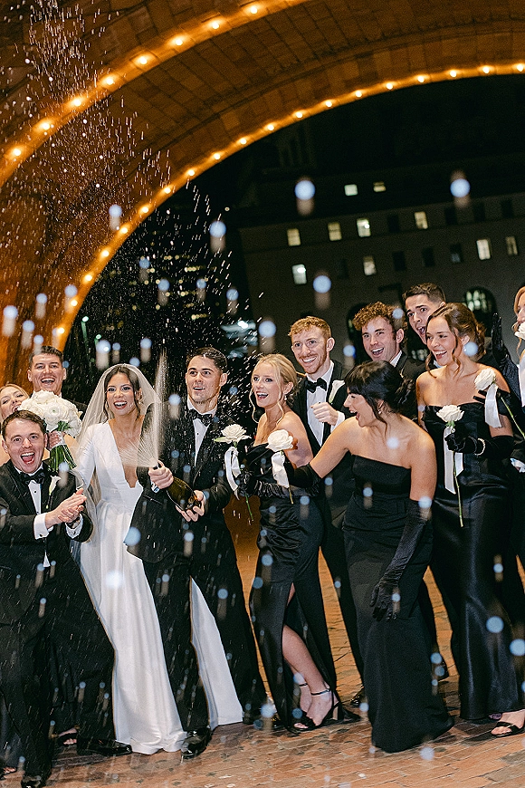 Wedding party celebration with champagne spray as bride in veil holds white rose bouquet, friends in tuxedos and black dresses under string lights on arched bridge at night