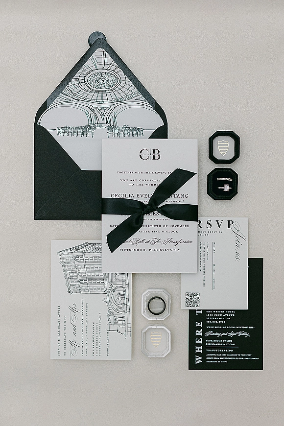 Invitation suite with black and white wedding invitations tied with a ribbon bow, wax seal, QR code RSVP, and rings on a neutral surface