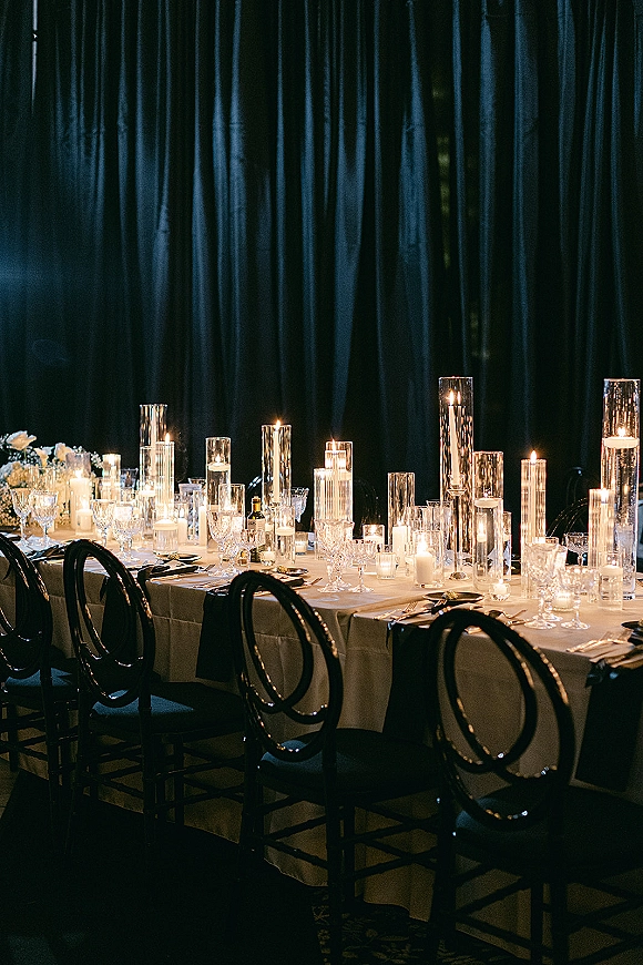 Reception tablescape with candlelit wedding table details, taper and hurricane vase candles, crystal glassware, floral centerpiece, dark draped backdrop