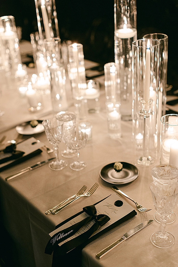 Reception tablescape with a candlelit wedding table featuring taper candles in glass vases, crystal goblets, black ribbon name cards and wax seal in a dark room