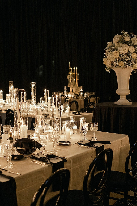 Reception tablescape with candlelit wedding reception details, black napkins on white linens, white rose urn centerpiece, black drape backdrop