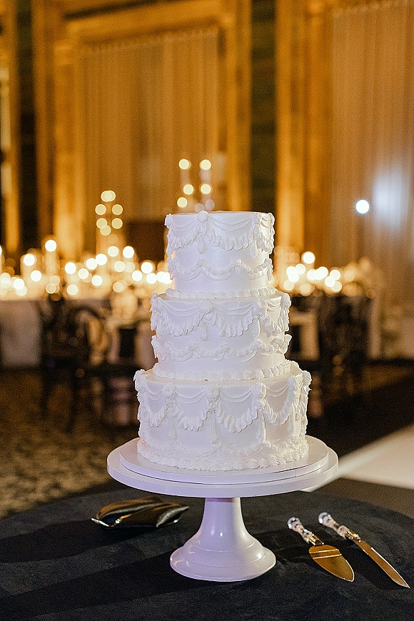Wedding cake three tier wedding cake with piped frosting garlands on a stand, set beside candles and a cake server in candlelit reception room