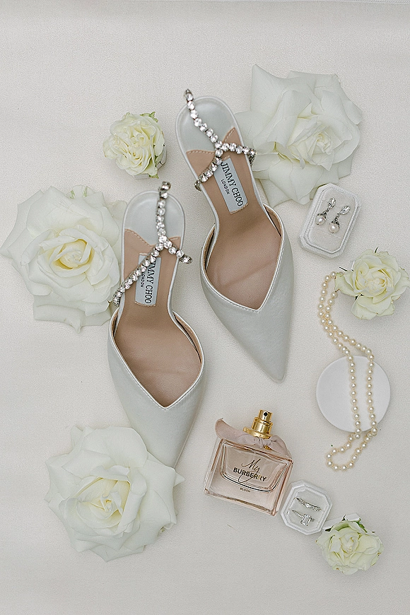 Bridal shoes flatlay featuring white pointed-toe heels with crystal straps, surrounded by white roses, pearls, ring and perfume on light fabric