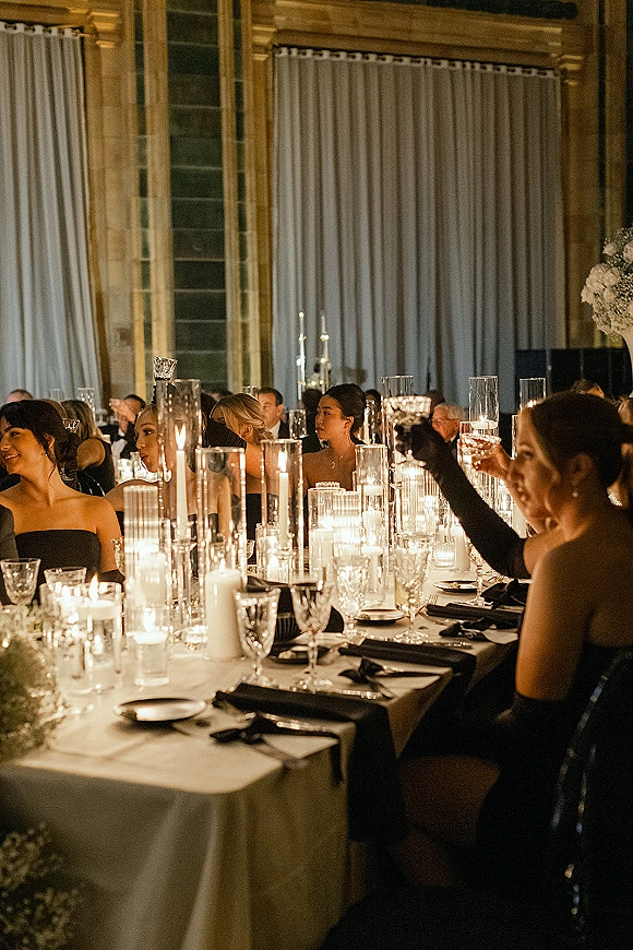 Reception tablescape with candlelit wedding reception glow, tall glass cylinder vases, white linens and black settings in a draped ballroom