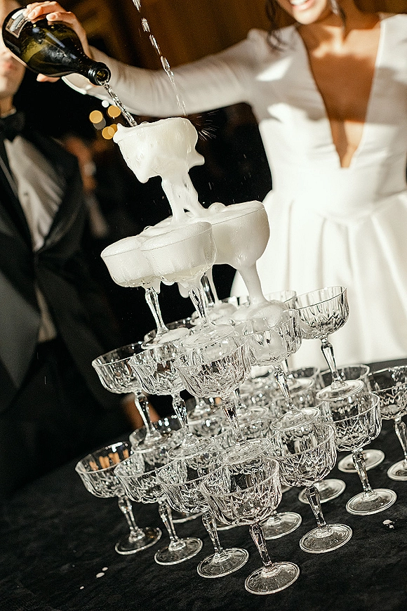 Champagne tower pour cascading through coupe glasses on a black tablecloth as a bride in a long sleeve dress pours under warm bistro lights