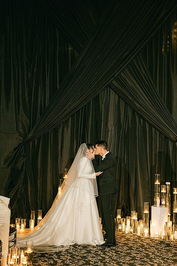 Wedding kiss portrait of bride in long sleeve gown and veil kissing groom in tuxedo beside pillar candles, against a dark draped backdrop