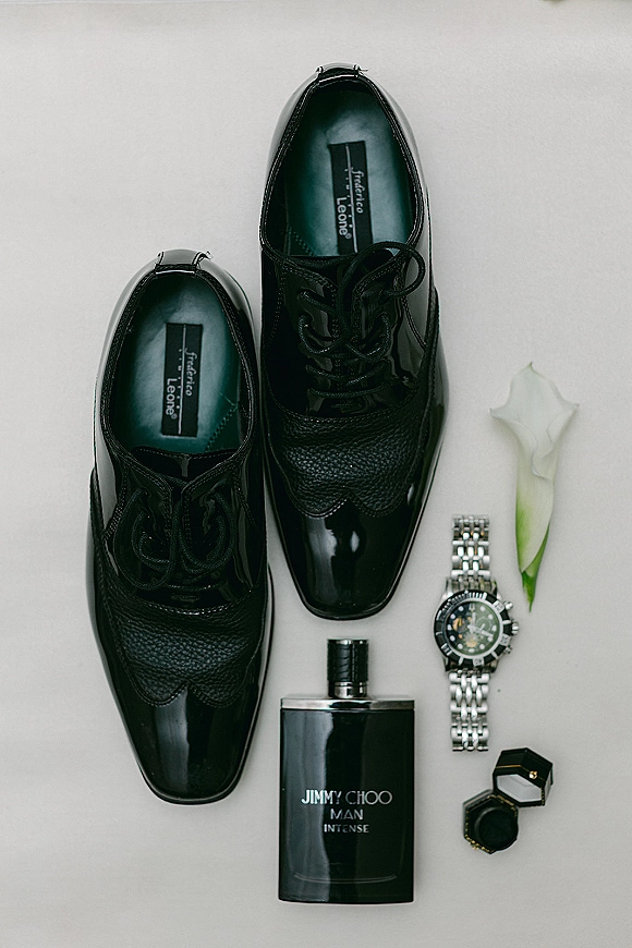Groom accessories flat lay with black dress shoes, wristwatch, cologne, ring box, and a calla lily on a light neutral surface