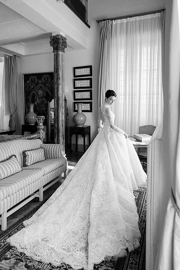 Bridal portrait in a long sleeve lace wedding dress with cathedral train, standing by large windows with sheer curtains in a living room