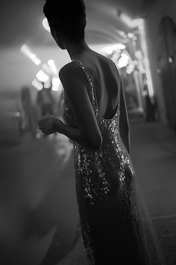 Wedding guest portrait in a sequin wedding guest dress with low back and updo, posed in an indoor corridor with blurred lights and guests silhouettes