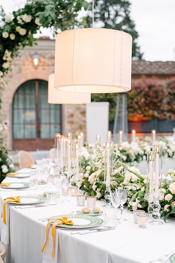 Reception tablescape with long wedding table setup featuring green plates, yellow ribbon napkins, taper candles, and lanterns on a stone patio