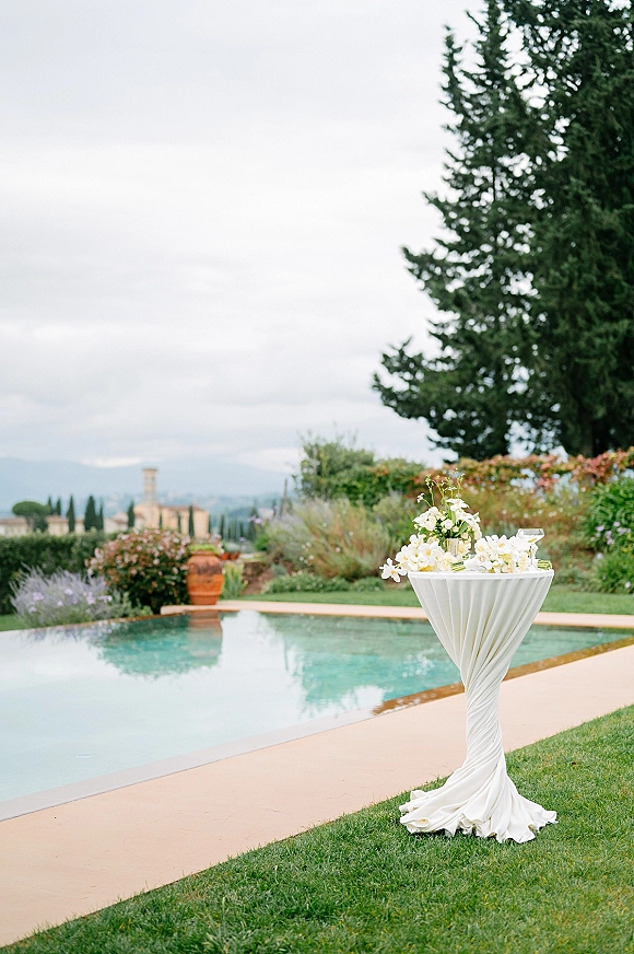 Wedding cocktail table with white linen and floral centerpiece, glassware set for poolside cocktail hour on a lawn with terracotta planters