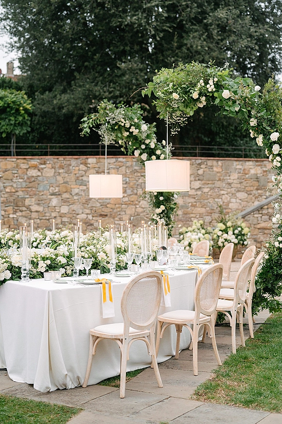 Reception tablescape with white floral garland, tall glass candlesticks, and yellow ribbon napkins beneath hanging lanterns by a stone wall
