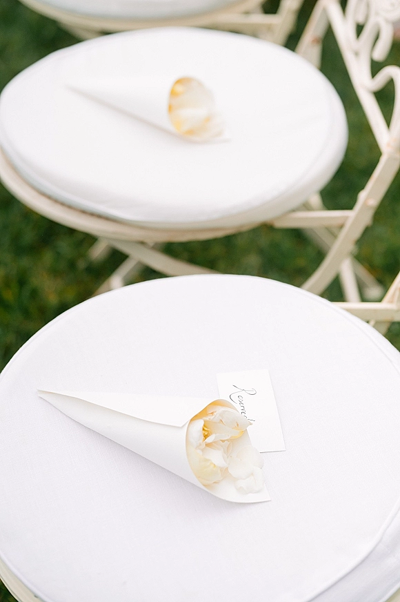 Ceremony chair decor with wedding petal cones, white cushions and escort card on a folding chair, petals scattered on grass lawn