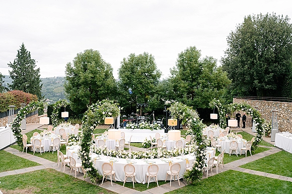 Outdoor reception setup with round white-linen tables, floral arches, greenery garlands, candles, and a live band stage on a lawn by trees