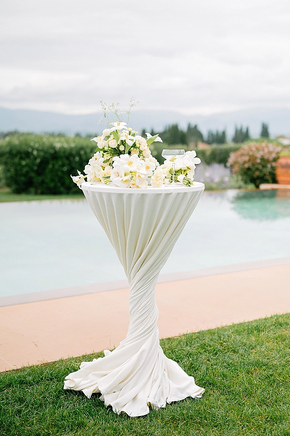 Cocktail table decor with a white orchid centerpiece and greenery on draped linen, set poolside on a lawn with distant hills under clouds