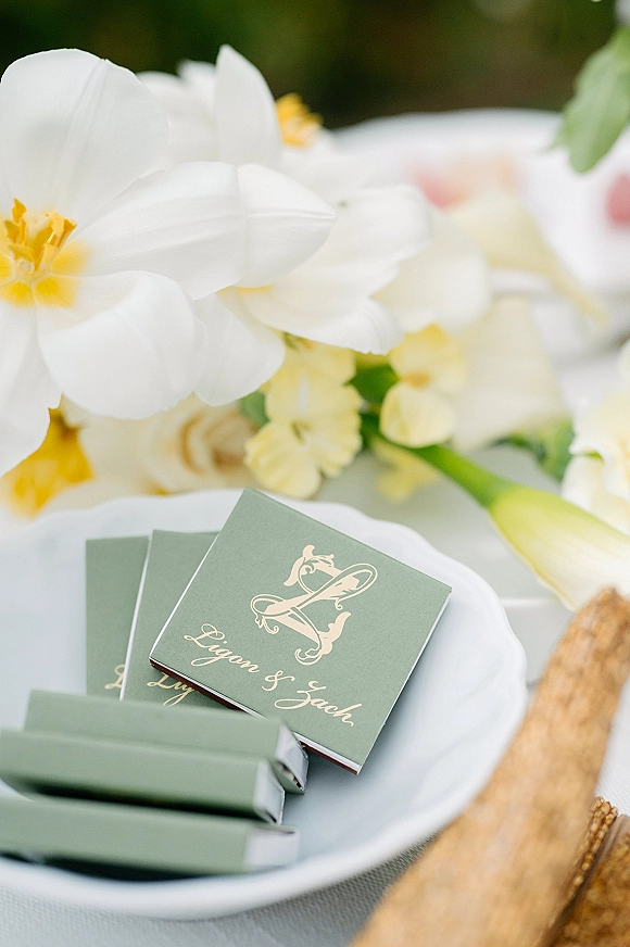 Wedding place cards in green with gold calligraphy and monogram, set on a white plate with napkin, wood charger, menu card, and white flowers