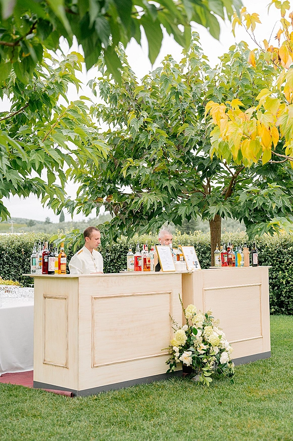 Wedding bar setup with outdoor wedding bar on a wood counter, liquor bottles, framed menu sign, glassware, and white hydrangeas under trees