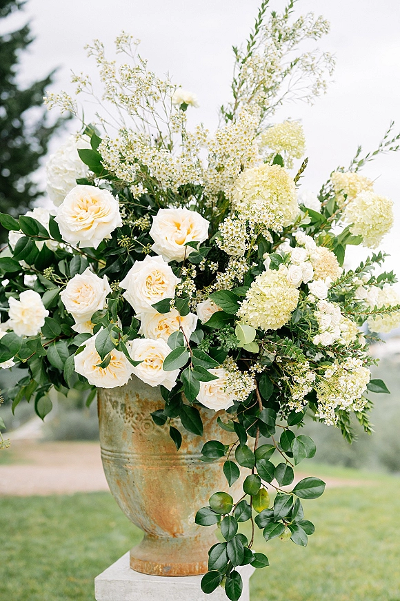 Wedding floral arrangement in a ceramic urn with white roses, hydrangea, baby’s breath, and greenery on an outdoor lawn with trees and sky