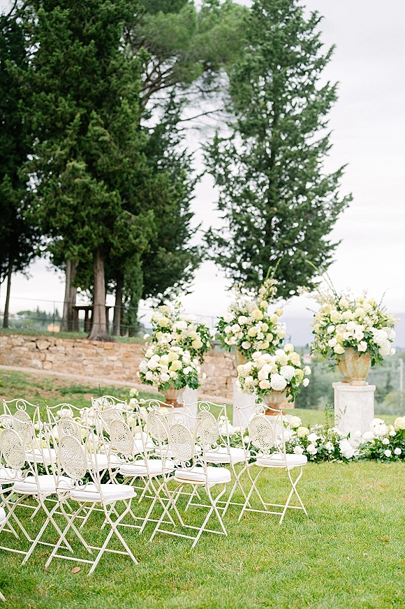Ceremony setup with white metal chairs and lush white-and-green floral arrangements lining an outdoor wedding ceremony lawn by a stone wall