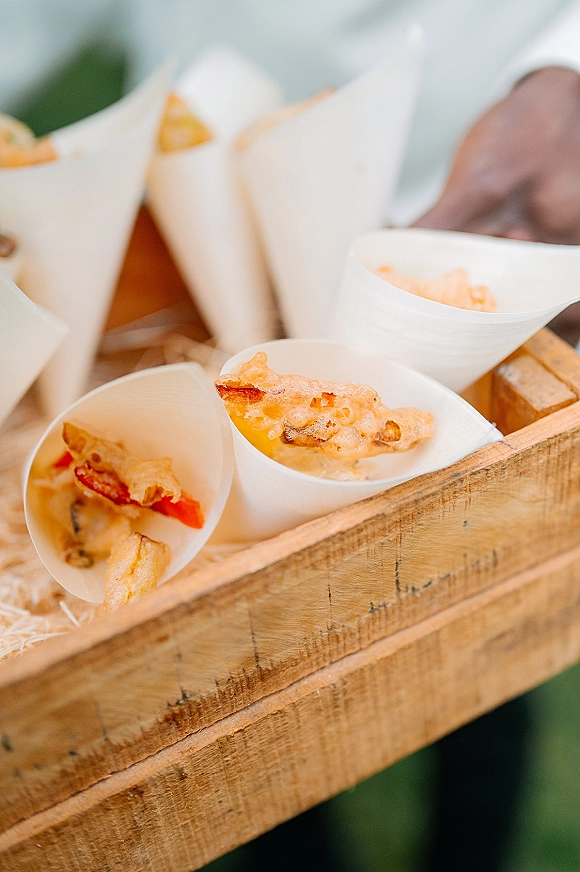 Wedding appetizers in paper food cones on a wooden serving tray, filled with fried bites against blurred greenery outdoors