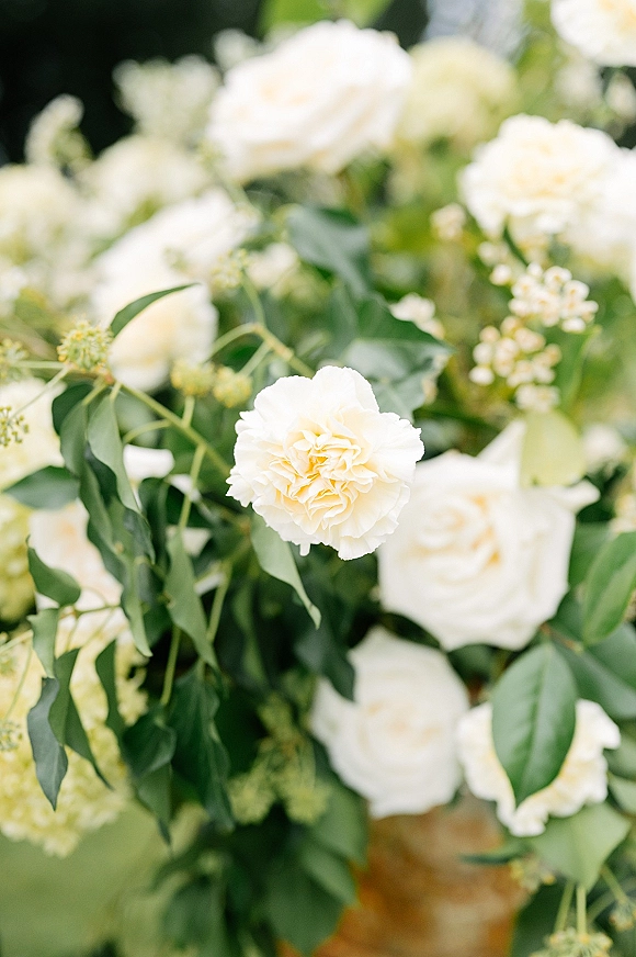 Wedding floral arrangement of white wedding flowers with roses, cream carnations, and lush greenery against a softly blurred garden backdrop