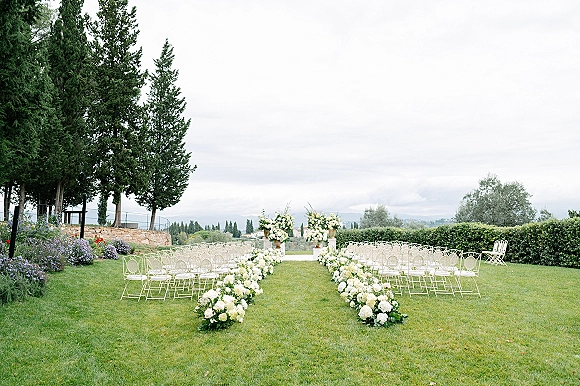 Outdoor ceremony setup with floral lined wedding aisle, white blooms and greenery by white chairs on a lawn with hedges and trees