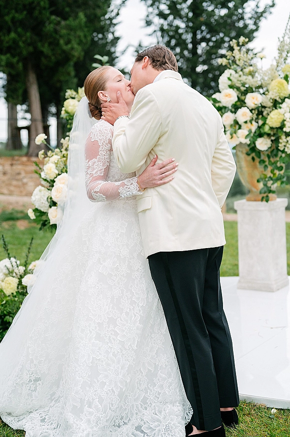 Wedding kiss under a white rose and greenery floral arch, bride in lace long-sleeve gown and veil, groom in white dinner jacket outdoors