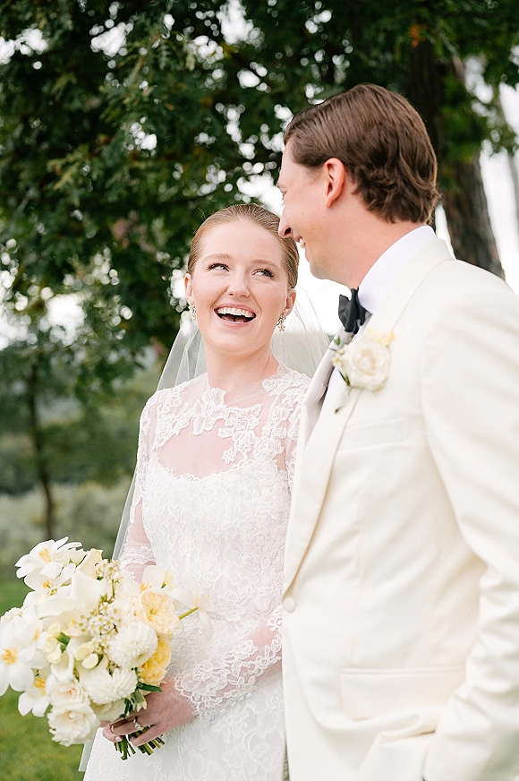 Couple portrait of bride laughing in a lace dress and veil holding a white and yellow bouquet as groom in cream tux watches in a garden setting