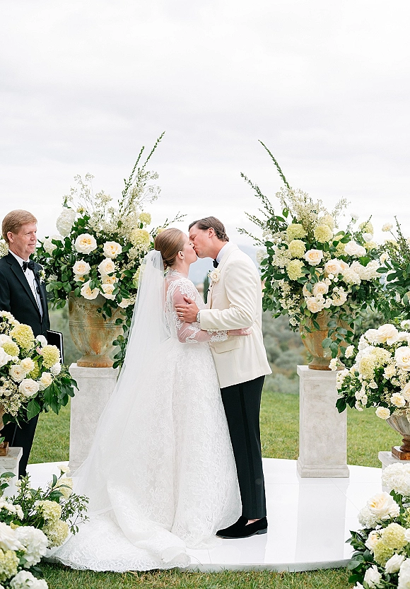 Wedding kiss at an outdoor wedding ceremony, bride in lace dress and veil with groom in white jacket, framed by urn florals on a lawn