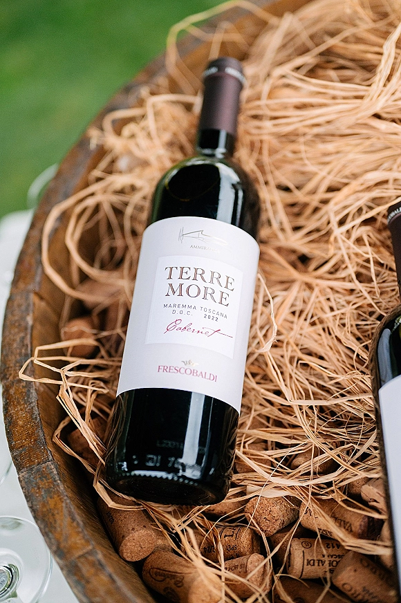 Wine bottle detail on a wooden barrel with corks and straw packing, beside wine glasses on a white table on a green lawn