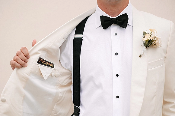 Groom suit detail with a white tuxedo jacket, black bow tie, and suspenders, finished with a rose boutonniere against a neutral wall