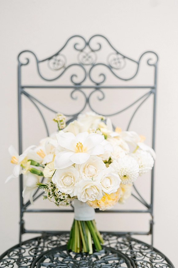 Bridal bouquet, white rose bridal bouquet with white blooms and yellow accents, wrapped stems resting on a wrought iron chair against a light wall