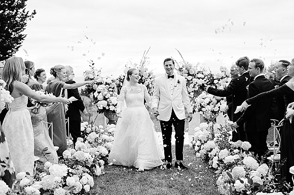 Wedding recessional as bride and groom walk hand in hand down a floral-lined outdoor aisle while guests toss flower petals under the sky