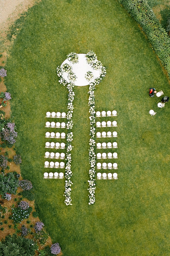 Ceremony setup with a white floral aisle leading to a round platform, white chairs arranged on a garden lawn with shrubs and flower beds