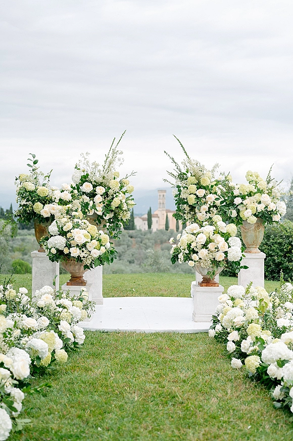 Ceremony altar decor with white roses and hydrangeas in large urns and ground florals on a white platform on a grassy estate lawn