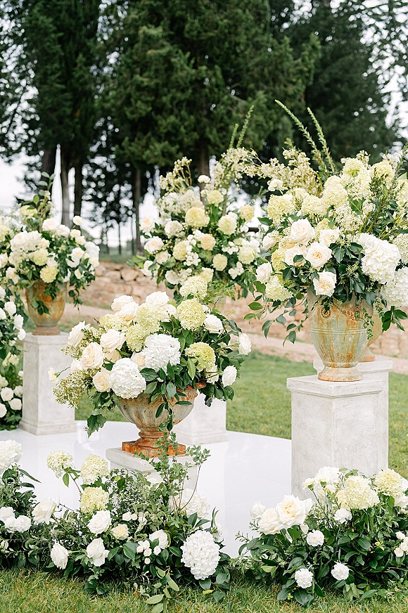 Ceremony floral decor with wedding aisle flowers in white roses and hydrangeas on stone pedestals, set on a lawn by tall trees and a stone wall