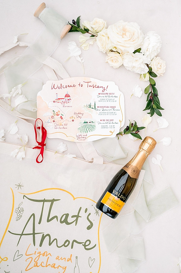 Wedding invitations with illustrated wedding invitation details, calligraphy lettering, red ribbon, white roses and petals beside a champagne bottle on white surface